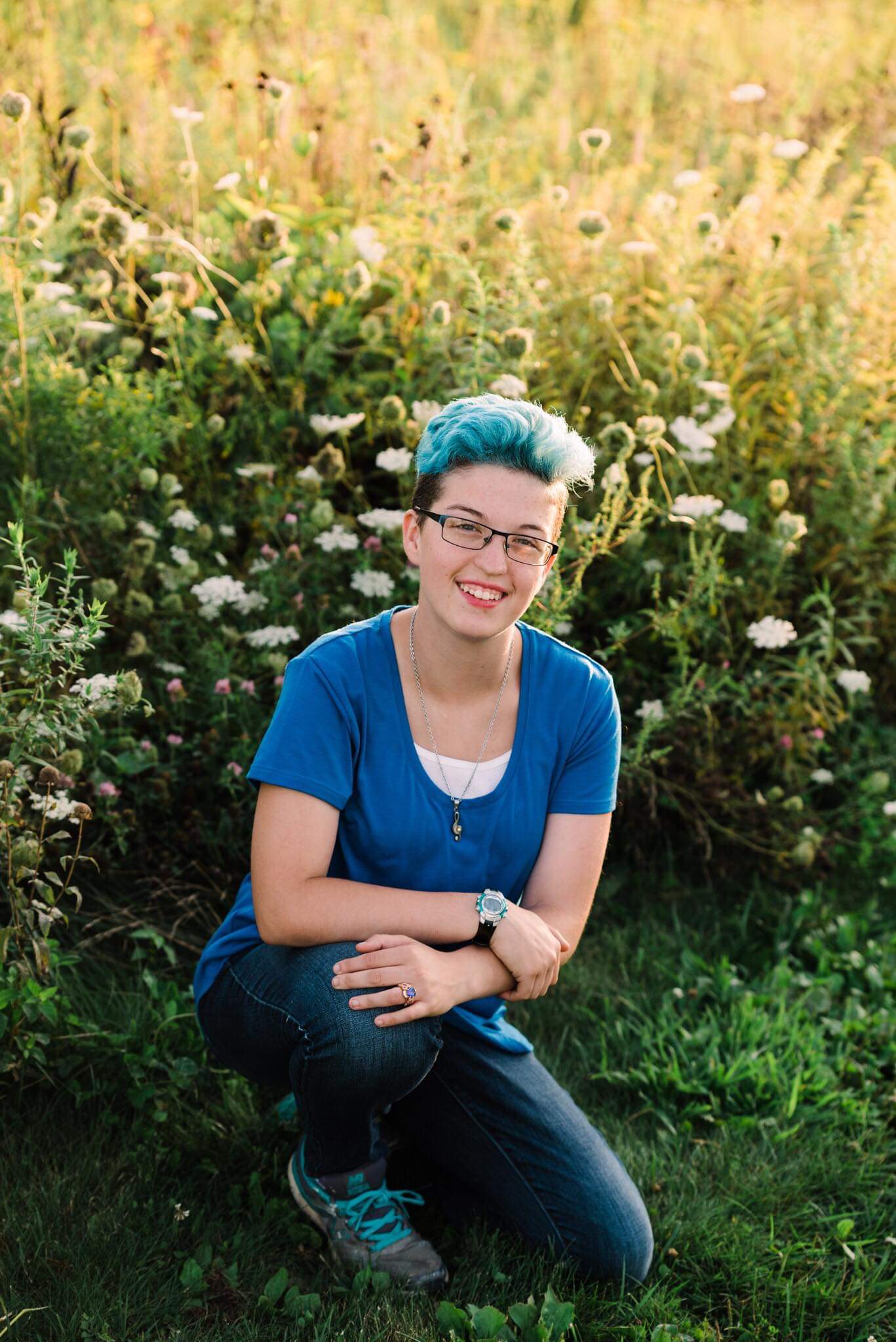 A young woman with blue hair, glasses, and a wristwatch wearing a predominantly blue business casual outfit. She is on one knee on a grassy path in a field of yellow wildflowers, posing for a professionally-taken high school senior photo.